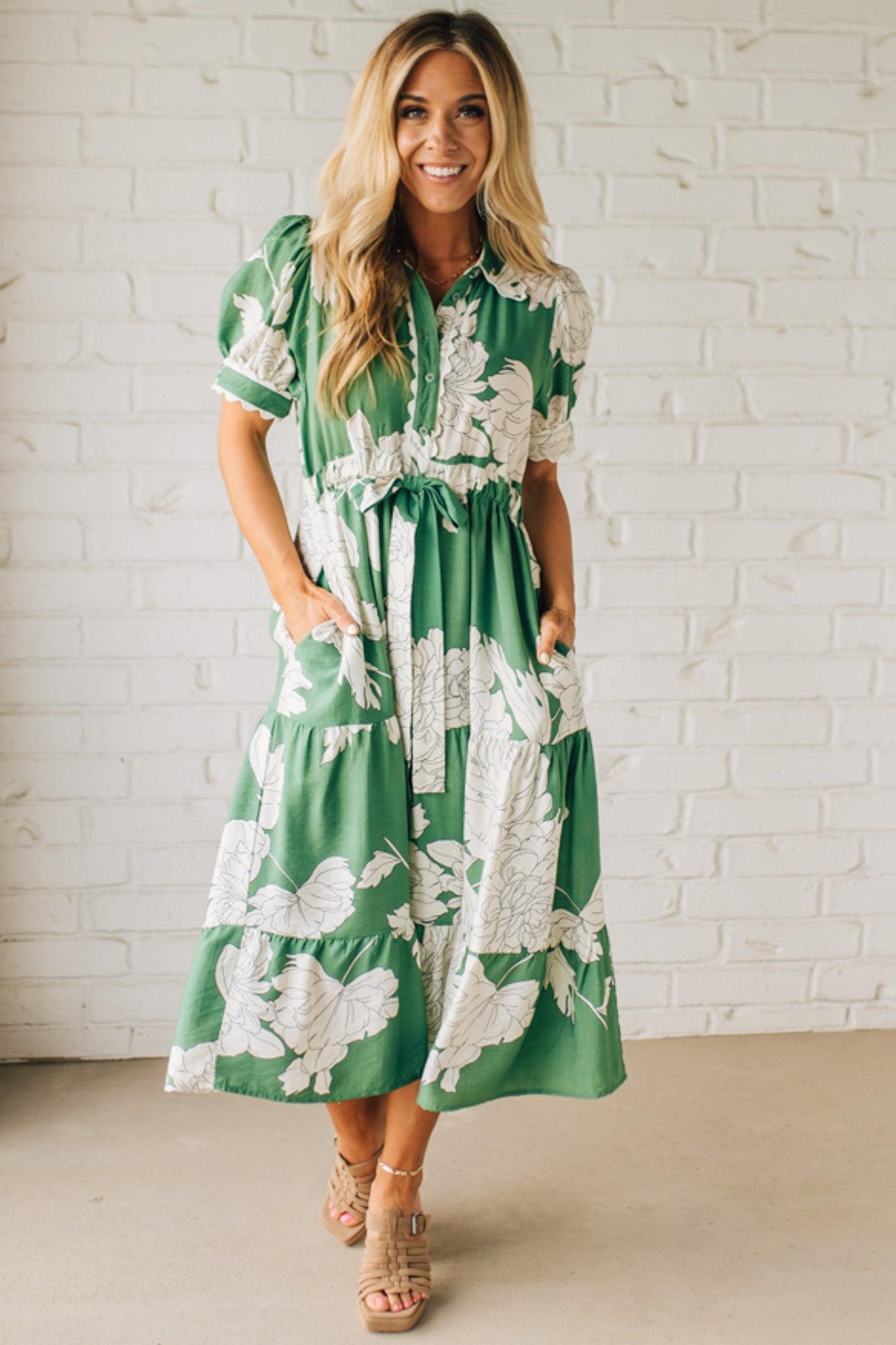 Blonde woman wearing a flower print midi dress with collared neck features buttoned closure, belted waistline, side pockets, puffed short sleeves, tiered layer skirt, and trimming along sleeves and neck.