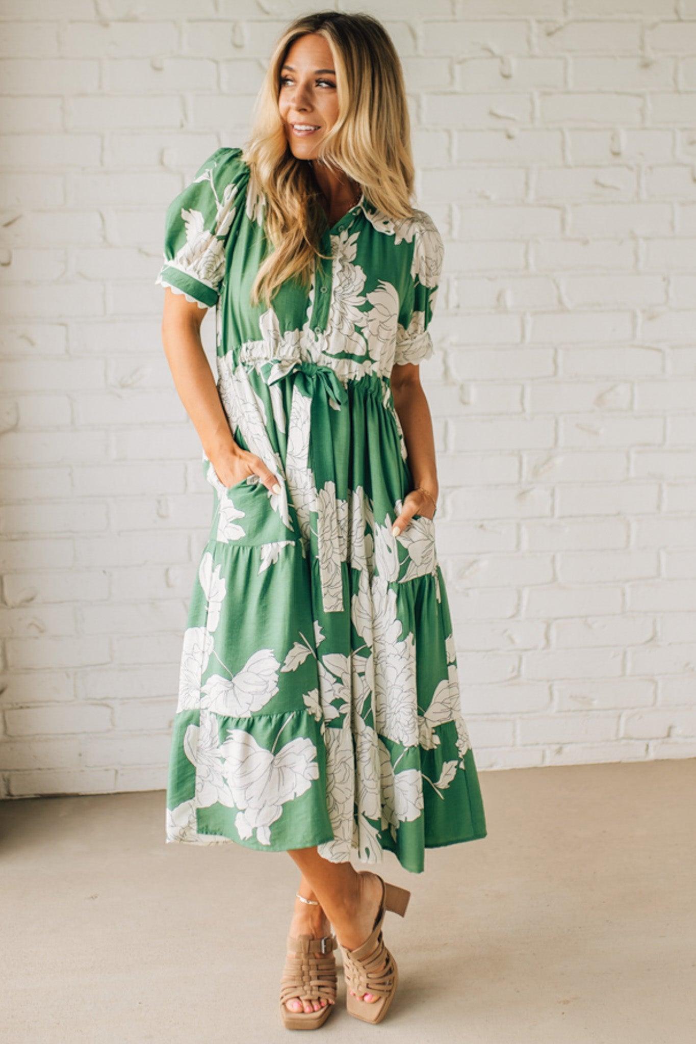 Blonde woman wearing a flower print midi dress with collared neck features buttoned closure, belted waistline, side pockets, puffed short sleeves, tiered layer skirt, and trimming along sleeves and neck.