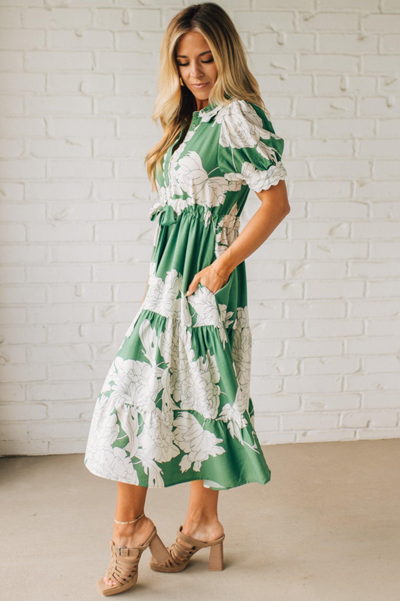 Blonde woman wearing a flower print midi dress with collared neck features buttoned closure, belted waistline, side pockets, puffed short sleeves, tiered layer skirt, and trimming along sleeves and neck.