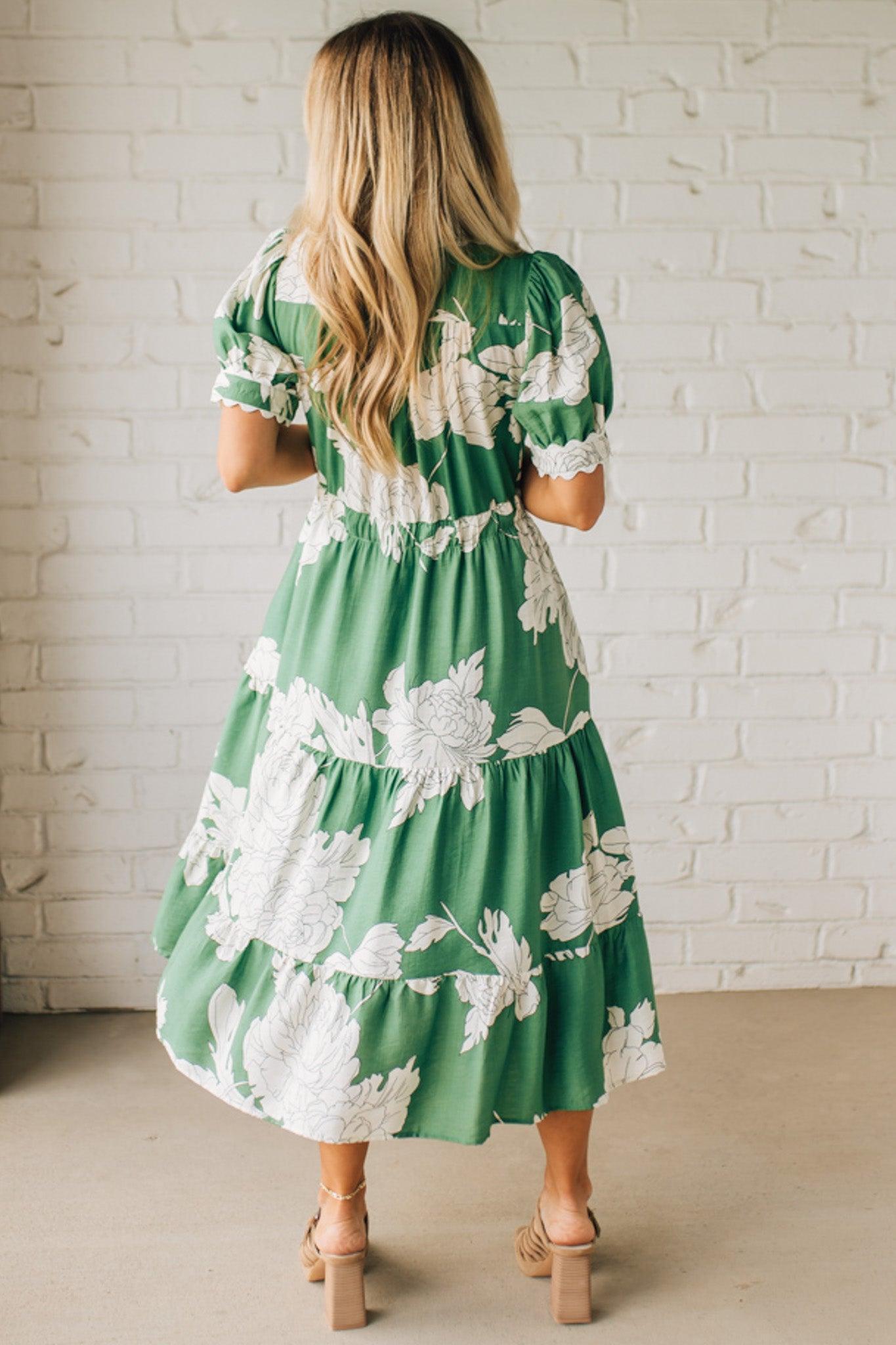 Blonde woman wearing a flower print midi dress with collared neck features buttoned closure, belted waistline, side pockets, puffed short sleeves, tiered layer skirt, and trimming along sleeves and neck.