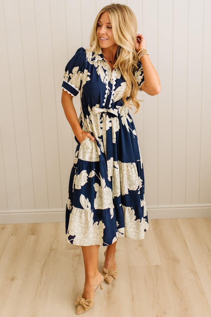 Woman wearing a navy and white floral dress against a light wooden paneled wall.