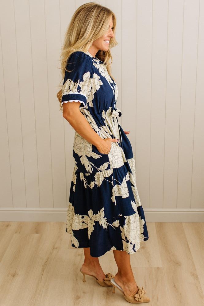 Woman wearing a navy and white floral dress against a light wooden paneled wall.