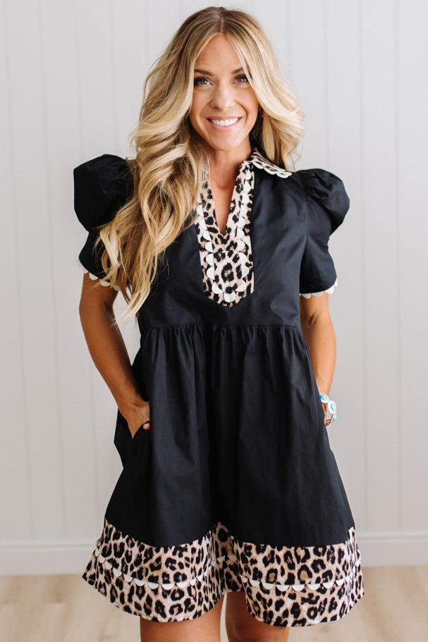 BLONDE WOMAN IN A BRIGHTLY LIT STUDIO WITH LIGHT WOOD FLOORS WEARING LIGHT TAN COWBOY BOOTS AND A BLACK KNEE LENGTH DRESS WITH LEOPARD PRINT ACCENT AND RICKRACK DETAILING.