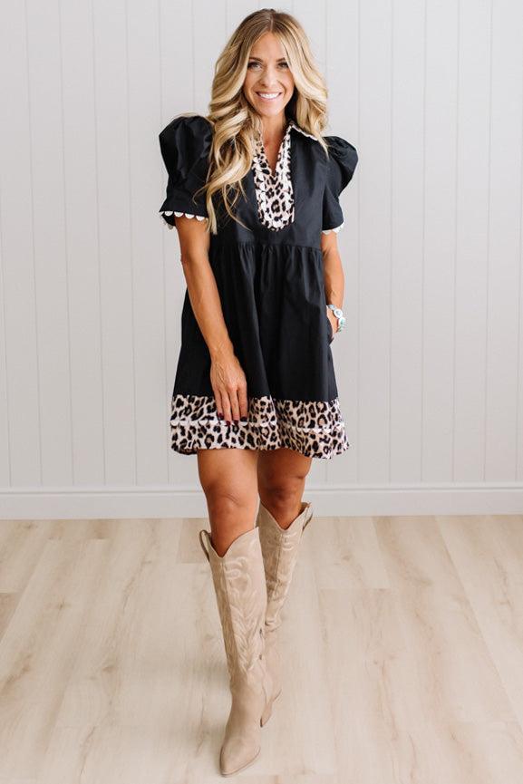 BLONDE WOMAN IN A BRIGHTLY LIT STUDIO WITH LIGHT WOOD FLOORS WEARING LIGHT TAN COWBOY BOOTS AND A BLACK KNEE LENGTH DRESS WITH LEOPARD PRINT ACCENT AND RICKRACK DETAILING.
