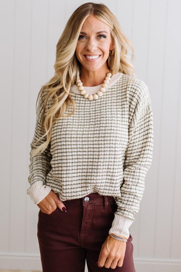 BLONDE WOMAN IN A BRIGHTLY LIT STUDIO WEARING A PAIR OF BROWN JEANS, CREAM CHUNKY BEADED NECKLACE AND A WAFFLED GRID SWEATER IN CREAM AND OLIVE GREEN.