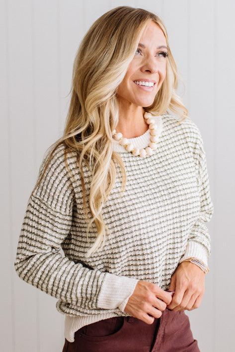 BLONDE WOMAN IN A BRIGHTLY LIT STUDIO WEARING A PAIR OF BROWN JEANS, CREAM CHUNKY BEADED NECKLACE AND A WAFFLED GRID SWEATER IN CREAM AND OLIVE GREEN.