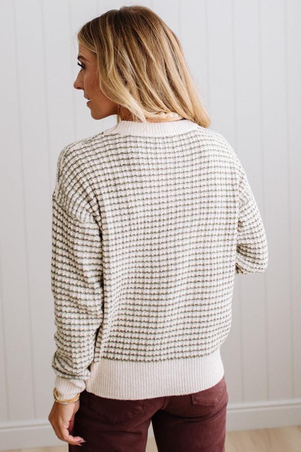 BLONDE WOMAN IN A BRIGHTLY LIT STUDIO WEARING A PAIR OF BROWN JEANS, CREAM CHUNKY BEADED NECKLACE AND A WAFFLED GRID SWEATER IN CREAM AND OLIVE GREEN.