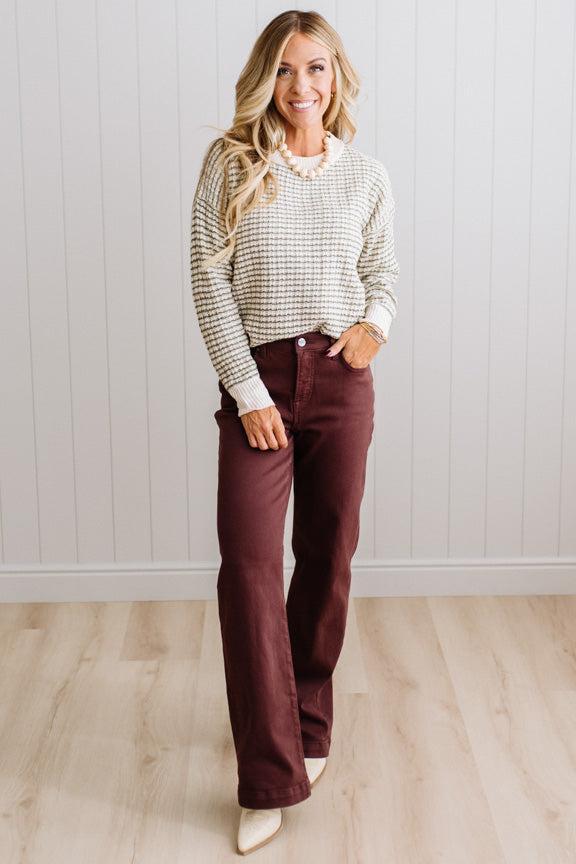 BLONDE WOMAN IN A BRIGHTLY LIT STUDIO WEARING A PAIR OF BROWN JEANS, CREAM CHUNKY BEADED NECKLACE AND A WAFFLED GRID SWEATER IN CREAM AND OLIVE GREEN.