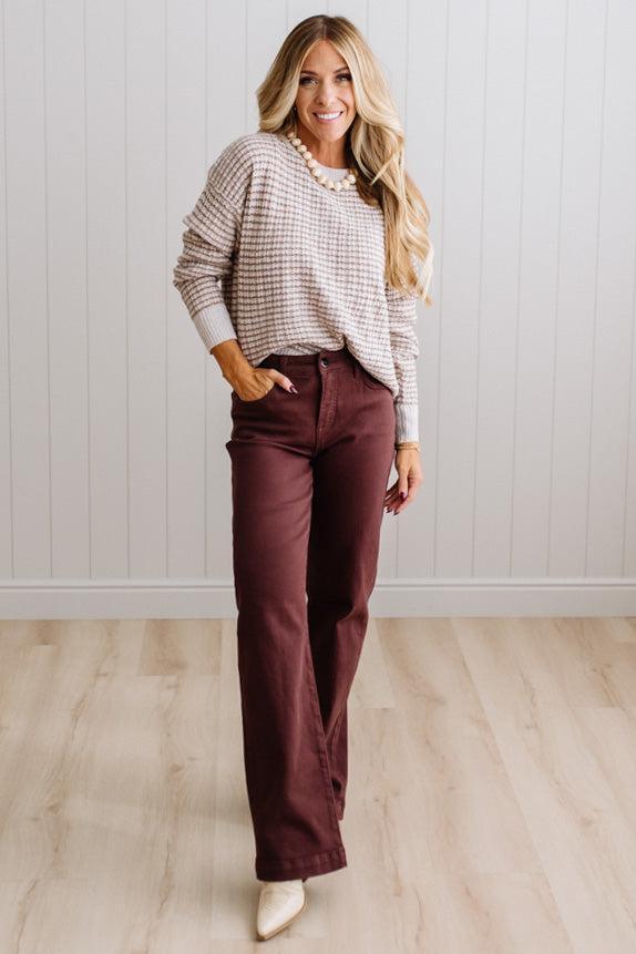 BLONDE WOMAN IN A BRIGHTLY LIT STUDIO WEARING A PAIR OF BROWN JEANS, CREAM CHUNKY BEADED NECKLACE AND A WAFFLED GRID SWEATER IN CREAM AND BROWN.