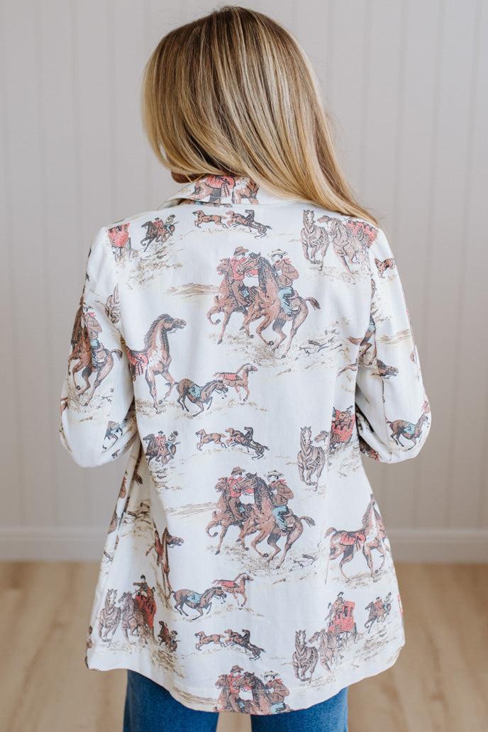 A woman viewed from behind wearing a white long-sleeve button-up shirt with a cowboy and horse print, paired with blue jeans, standing in front of a light wood-paneled wall.