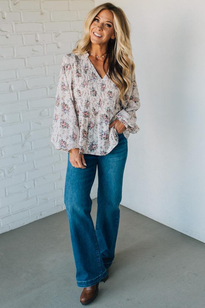 Woman wearing a floral blouse and blue jeans standing against a white brick wall.