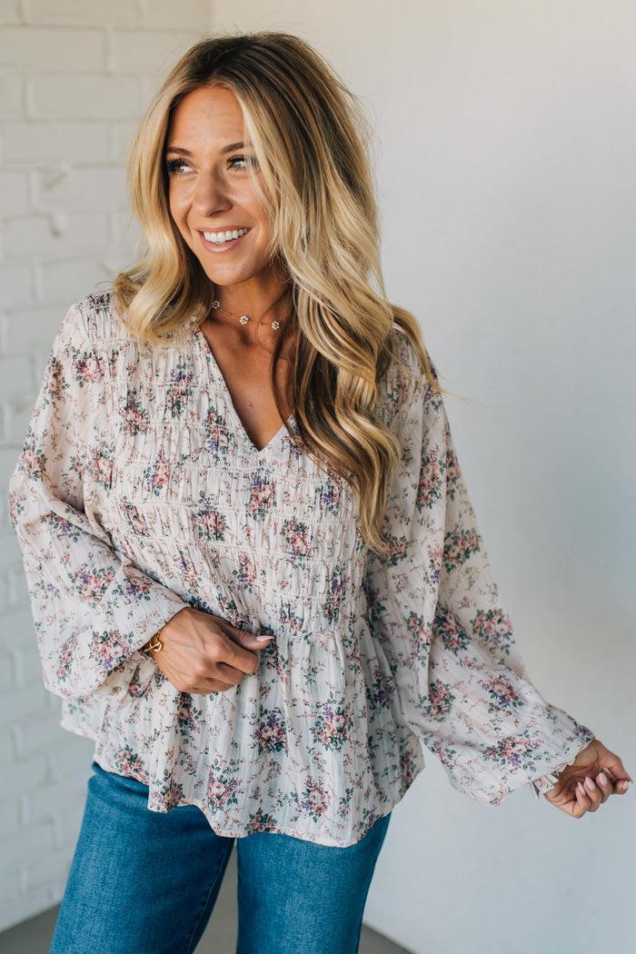 Woman wearing a floral blouse and blue jeans against a white brick wall.