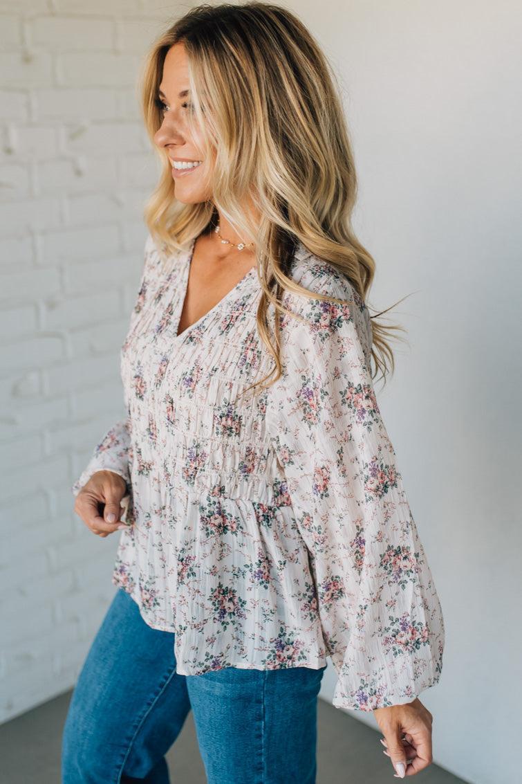 Woman wearing a floral blouse and blue jeans against a white brick wall.