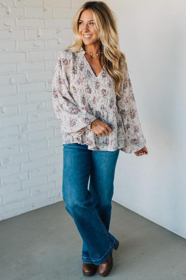Woman wearing a floral blouse and blue jeans against a white brick wall.