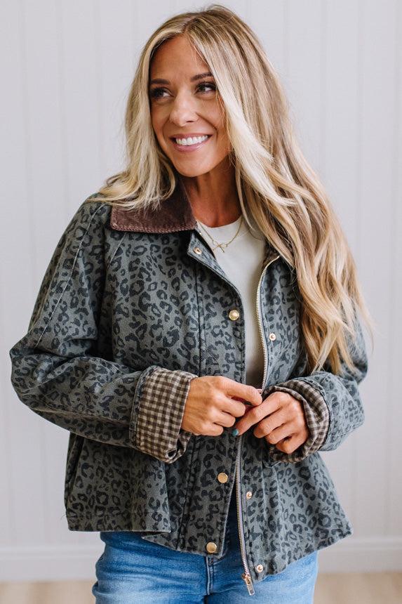 A woman wearing a dark gray leopard-print jacket with a brown corduroy collar and gold-tone buttons, paired with a beige top and blue jeans, standing against a white paneled wall.
