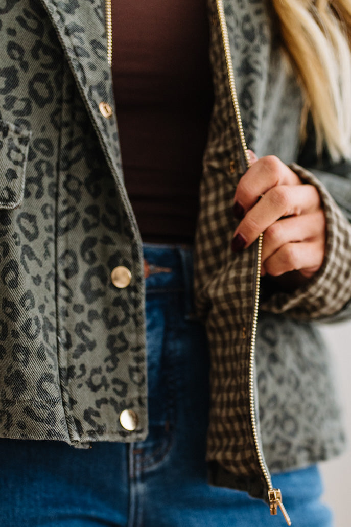 Close-up of a person wearing a cropped olive green leopard-print jacket with gold-tone snap buttons and a gold zipper, layered over a dark brown top and blue jeans.