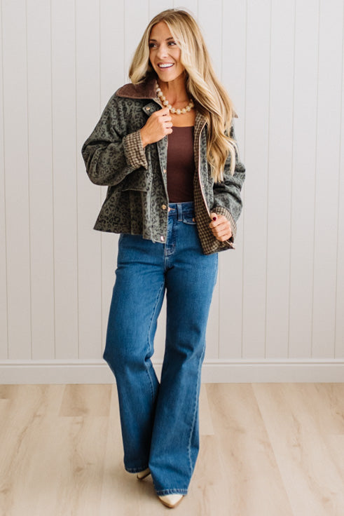 A woman modeling a cropped patterned jacket with a brown collar, worn over a brown top and blue flare jeans, standing against a white paneled wall and light wood floor.