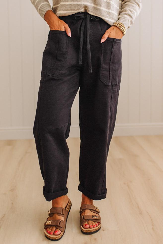 A person wearing dark charcoal cargo-style pants with a drawstring waist, shown from the waist down in a home setting with light wood flooring and paneled walls.