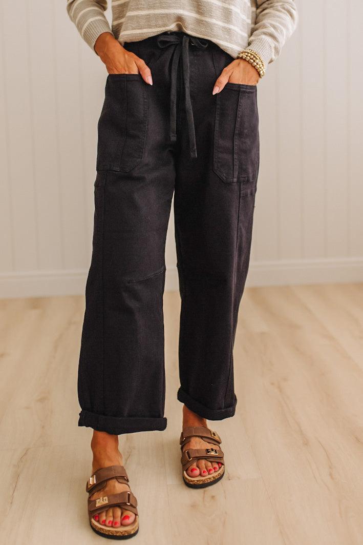 A person wearing loose black wide-leg pants with a drawstring waist, large front patch pockets, and rolled cuffs, paired with brown slide sandals and a striped long-sleeve top, standing on a light wood floor against a white paneled wall.