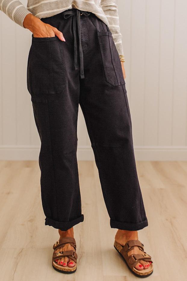 A person wearing loose black wide-leg pants with a drawstring waist, side pockets, and cuffed hems, paired with brown sandals and a light striped top, standing on a light wood floor against a white paneled wall.