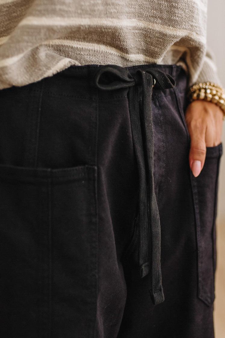 Close-up of a person wearing black drawstring pants with side pockets, paired with a beige striped knit top and a gold beaded bracelet.