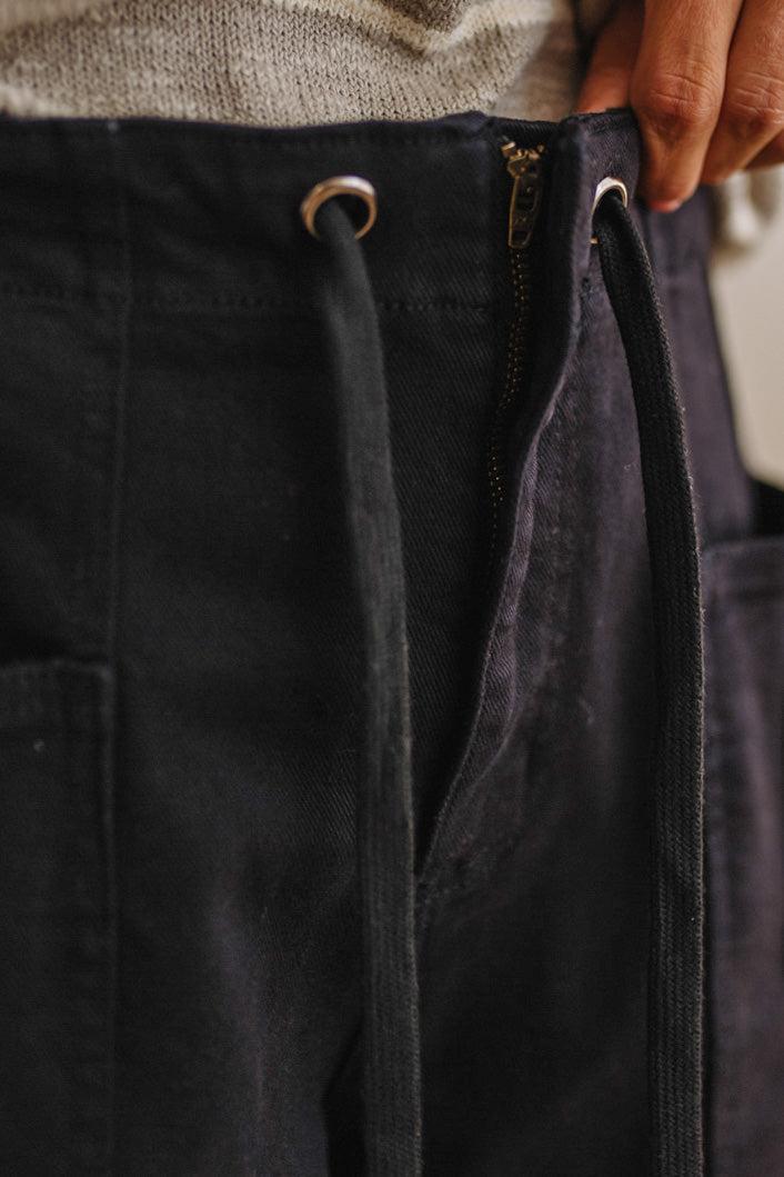 Close-up of dark charcoal gray pants with a drawstring waist and zipper closure, worn with a light gray knit top, with a hand adjusting the waistband.