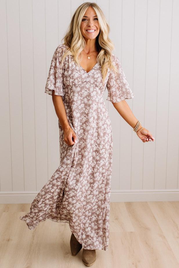 A woman modeling a loose, knee-length floral dress with short flutter sleeves and a V-neckline, standing in a room with light wood floors and a white paneled wall.