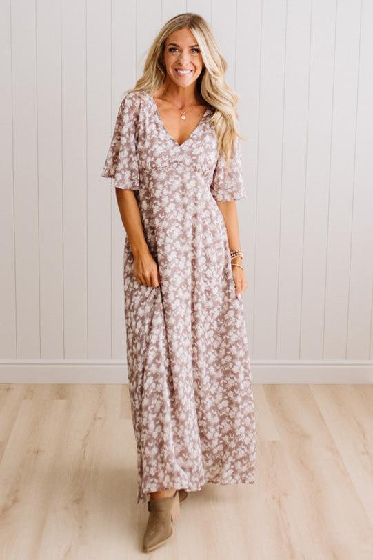 A woman modeling a long-sleeve, V-neck maxi dress with a dusty rose and white floral pattern, standing in front of a white paneled wall and light wood floor.