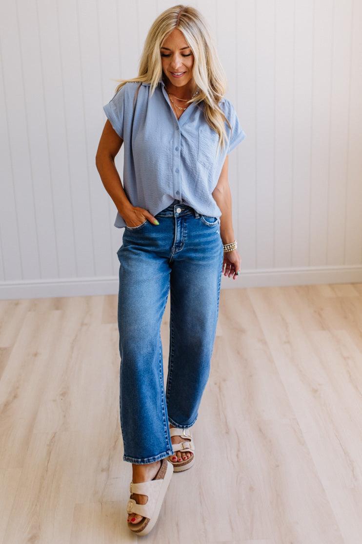 Woman wearing a light blue button-up shirt and jeans standing on a wooden floor.