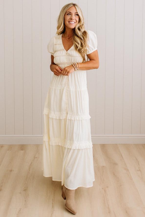 A woman modeling a cream-colored, tiered maxi dress with short puff sleeves and a V-neckline, standing in front of a white paneled wall and light wood floor.