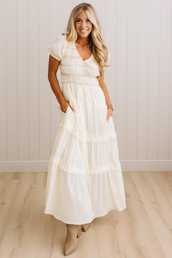 A woman modeling a cream-colored, tiered maxi dress with ruffled details and short flutter sleeves, standing in a room with light wood floors and white paneled walls.