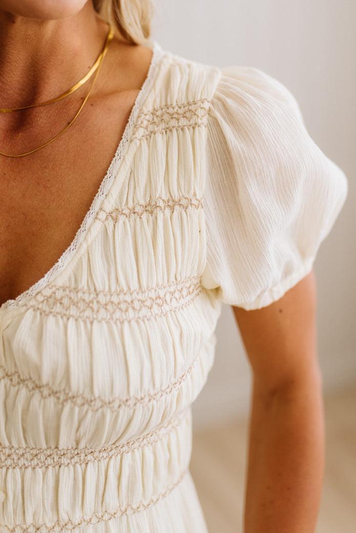 Close-up of a cream-colored smocked dress with short puff sleeves and delicate gold chain necklaces worn by a model.