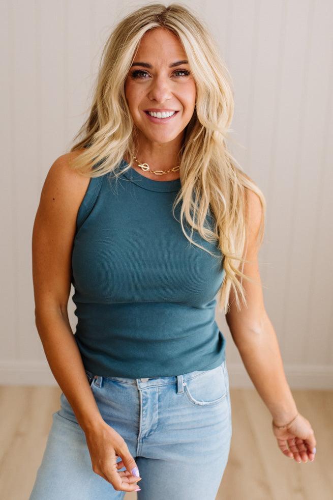 Woman wearing a teal sleeveless top and light blue jeans, standing against a plain background.