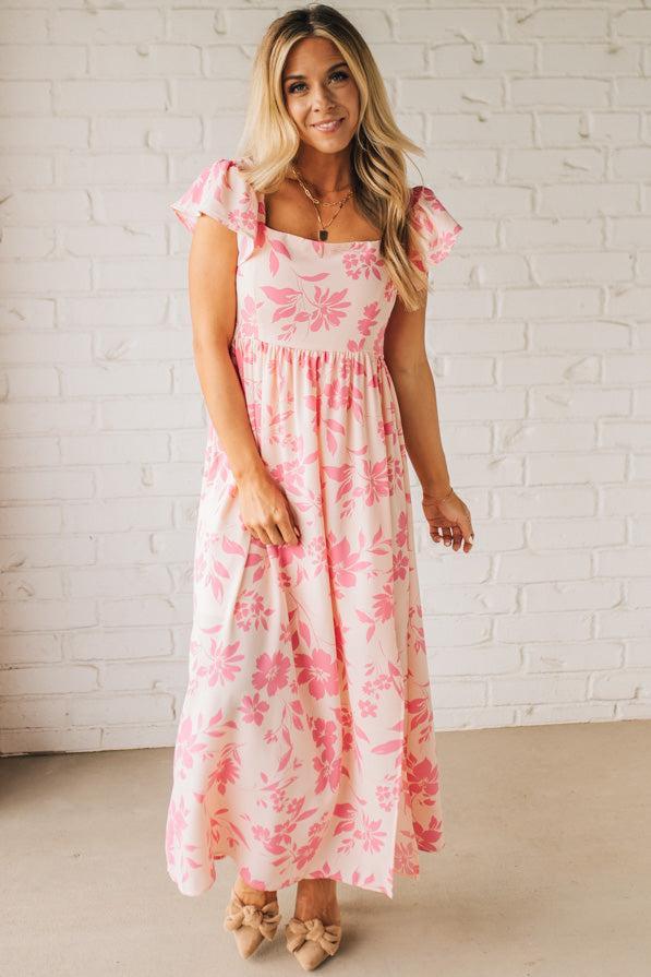 Blonde woman wearing a maxi dress with floral pink and a dark pink print, ruffle sleeved, with back smocked panel.