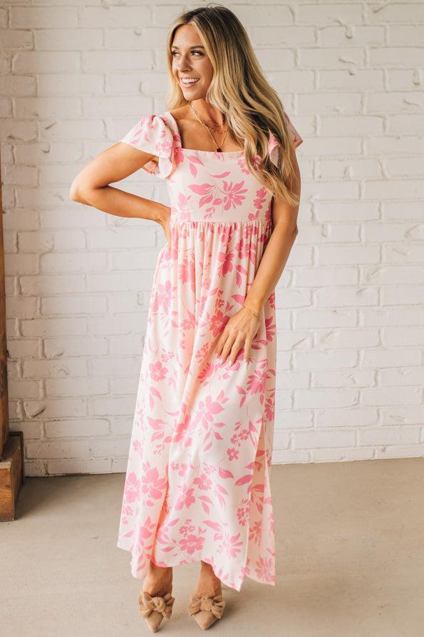 Blonde woman wearing a maxi dress with floral pink and a dark pink print, ruffle sleeved, with back smocked panel.