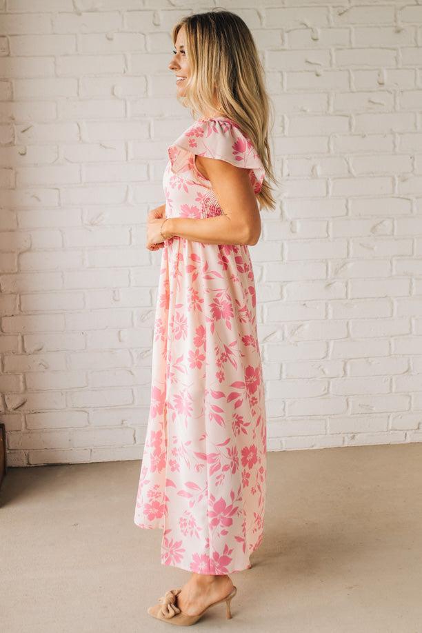 Blonde woman wearing a maxi dress with floral pink and a dark pink print, ruffle sleeved, with back smocked panel.