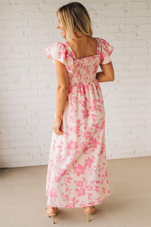 Blonde woman wearing a maxi dress with floral pink and a dark pink print, ruffle sleeved, with back smocked panel.
