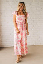 Blonde woman wearing a maxi dress with floral pink and a dark pink print, ruffle sleeved, with back smocked panel.