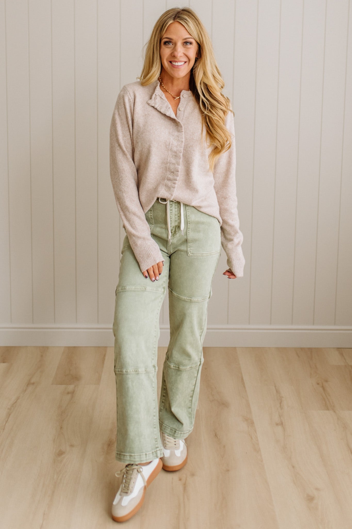 Woman wearing a light pink shirt and green pants standing against a white paneled wall.
