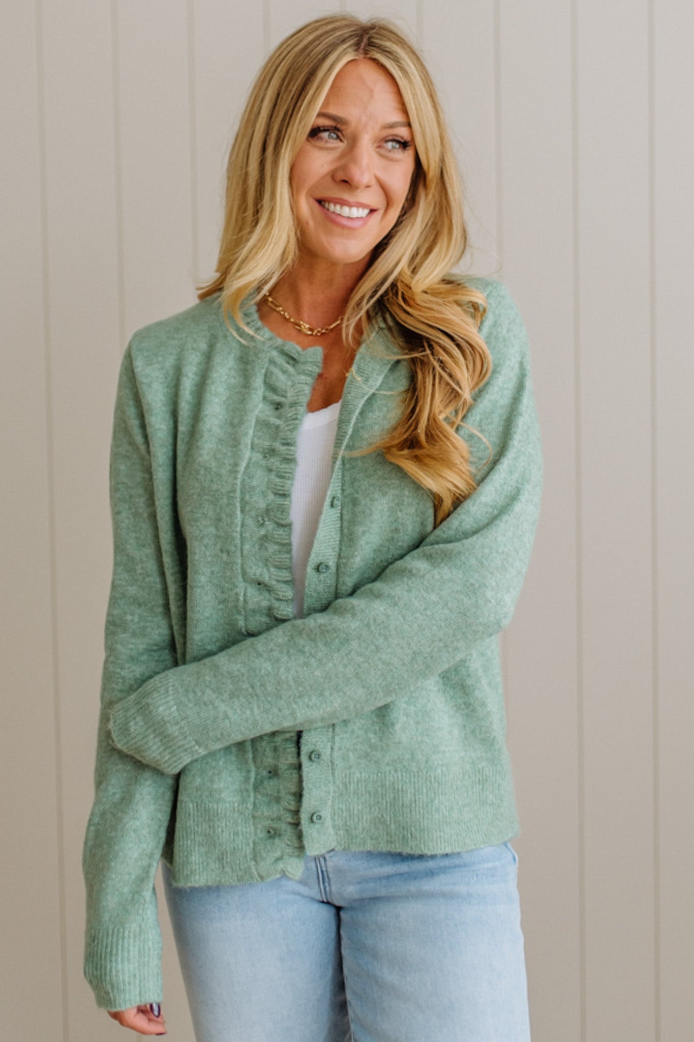 Woman wearing a green cardigan over a white shirt and blue jeans against a beige wall.