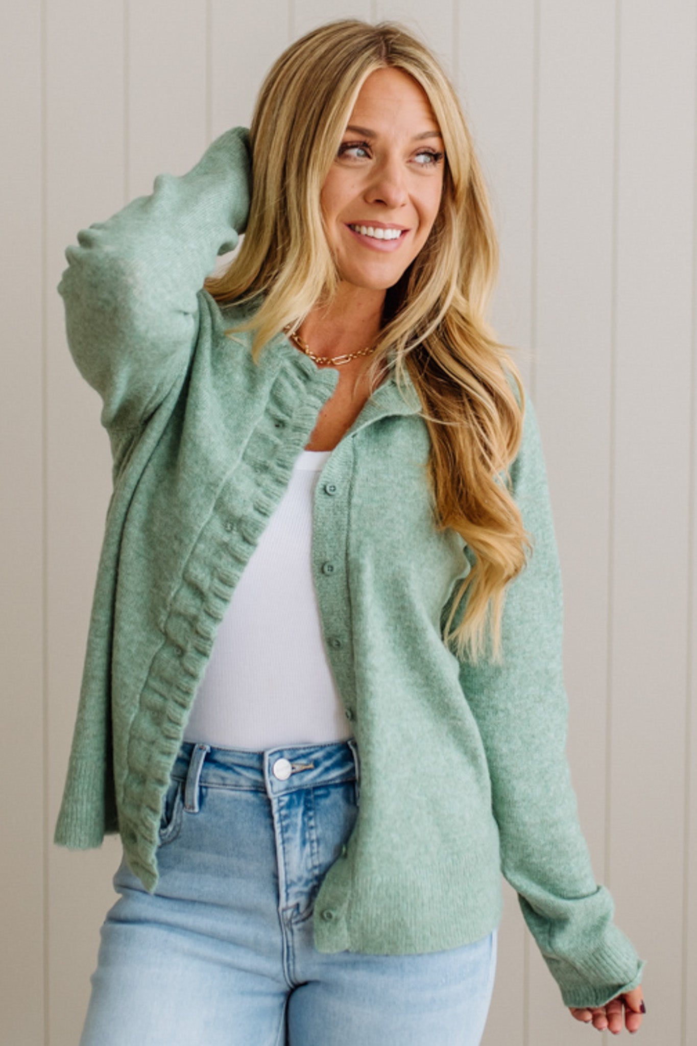 Woman wearing a green cardigan over a white shirt and blue jeans, standing against a light wooden panel background.