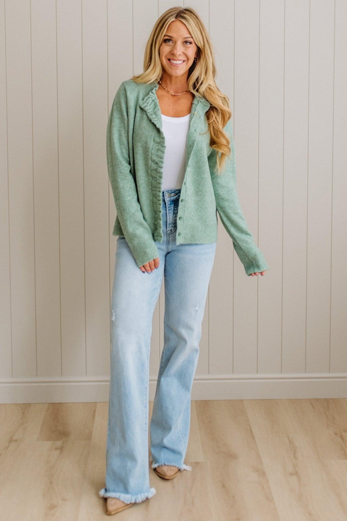 Woman wearing a green jacket, white top, and light blue jeans against a beige wall.