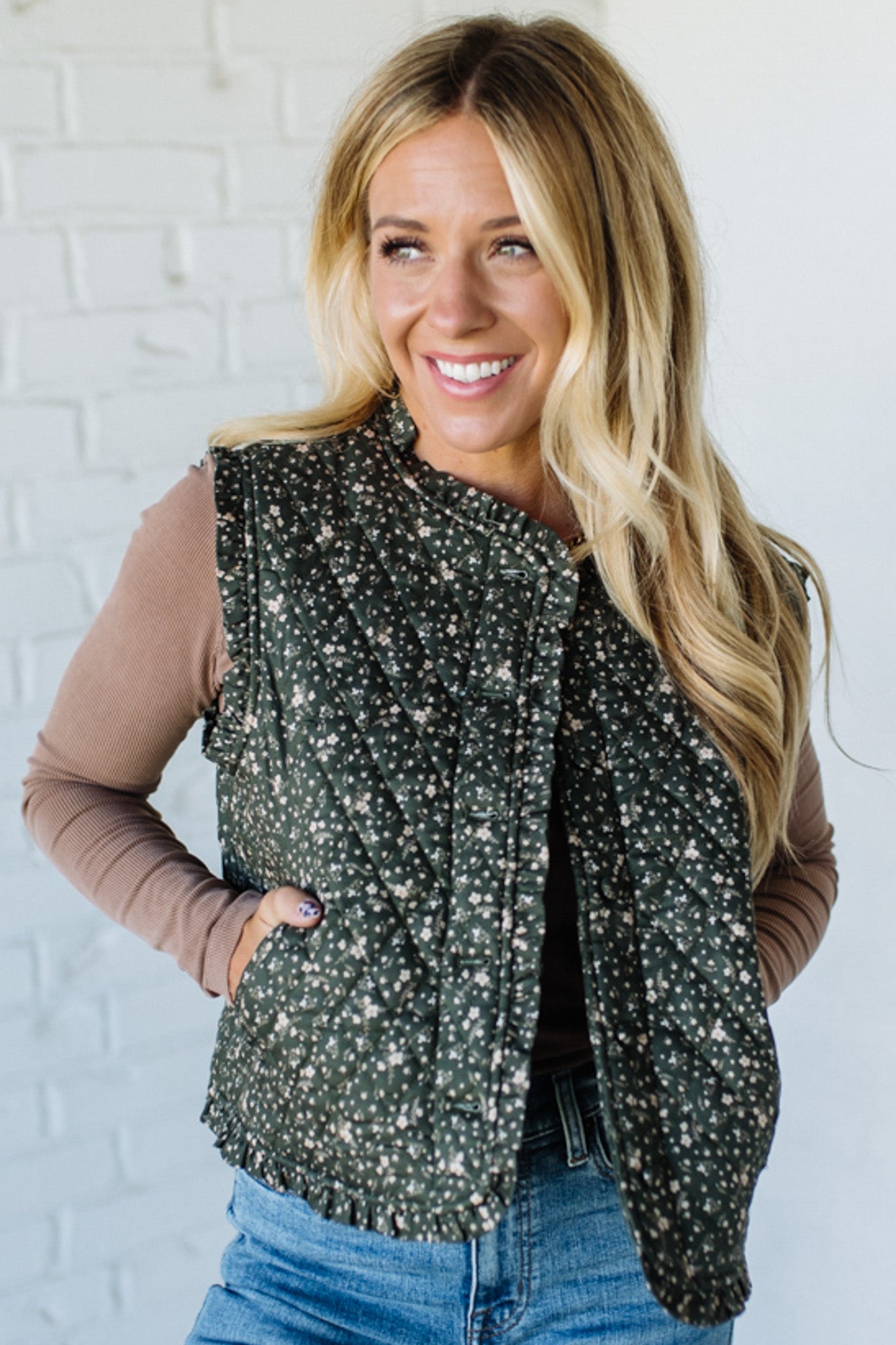 Woman wearing a floral green vest over a brown top and blue jeans against a white brick wall.