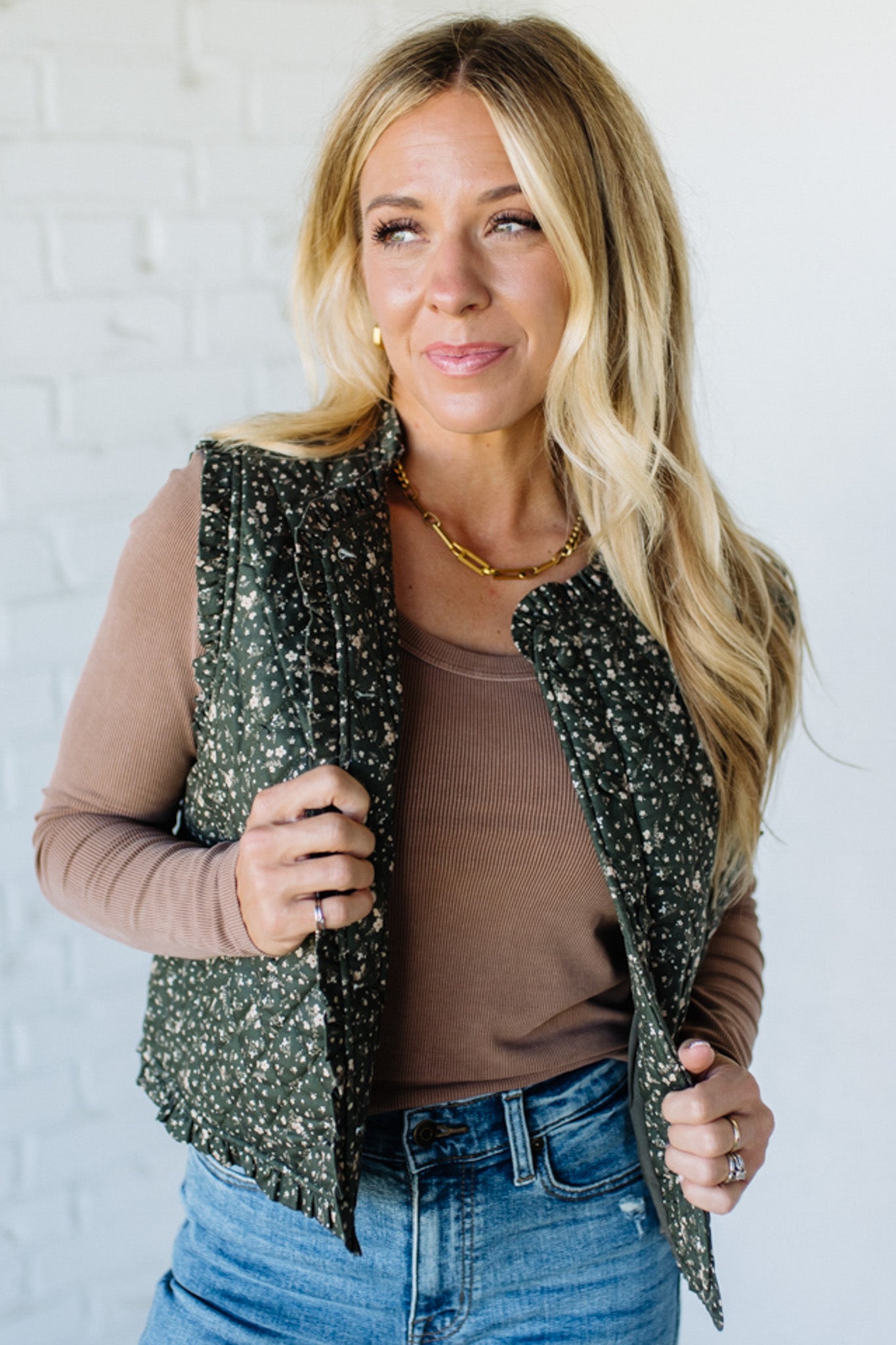 Woman wearing a floral green vest over a brown top and blue jeans against a white brick wall.