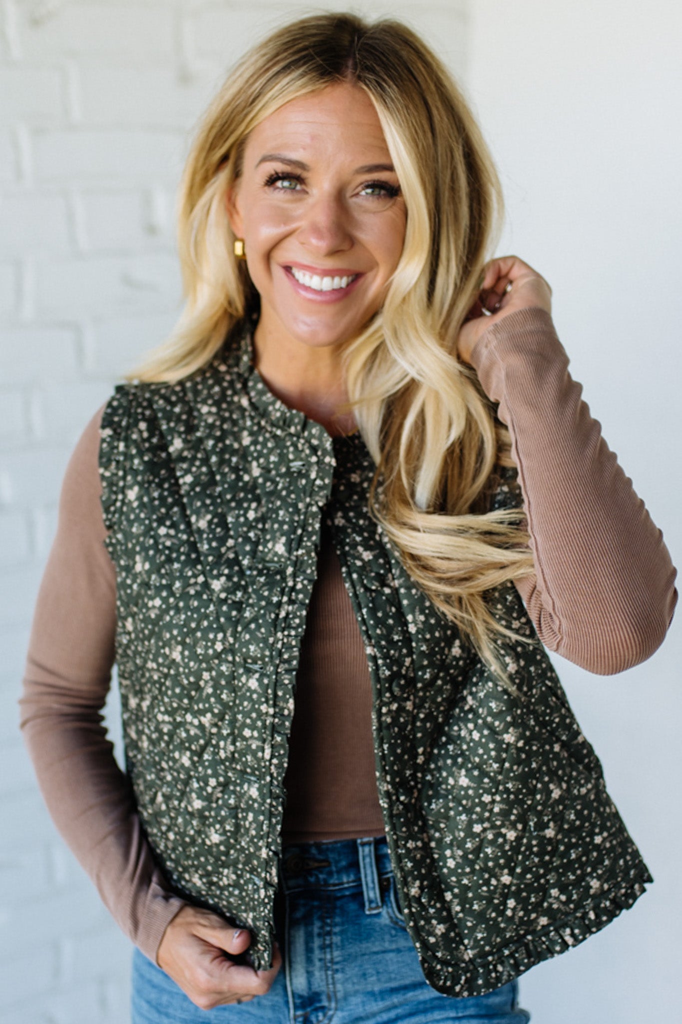 Woman wearing a floral green vest over a brown top and blue jeans against a white brick wall.