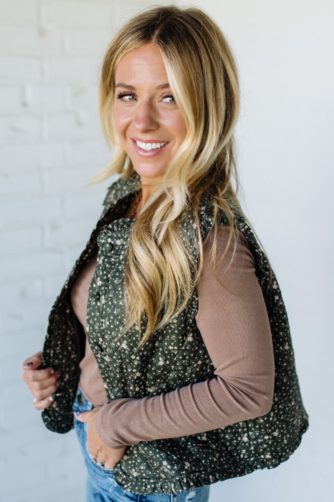 Woman wearing a floral green vest over a brown top and blue jeans against a white brick wall.