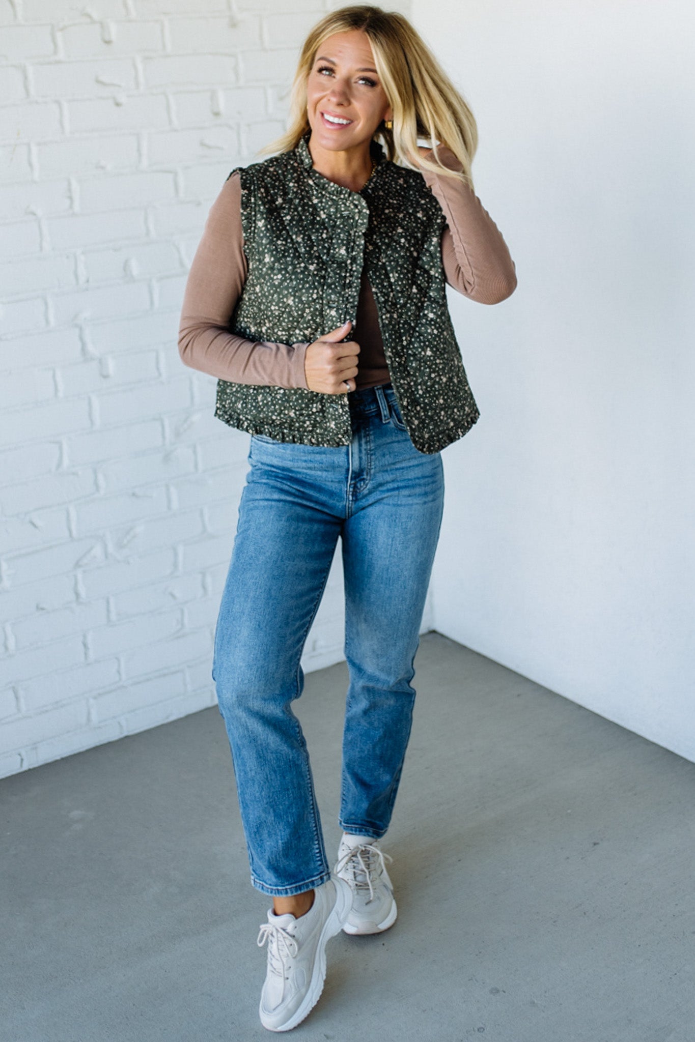 Woman wearing a floral green vest over a brown top and blue jeans against a white brick wall.