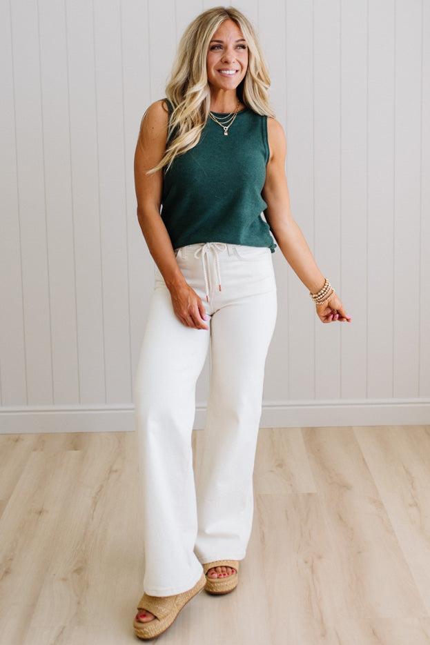Woman wearing a green sleeveless top and white pants standing against a light wooden paneled wall.