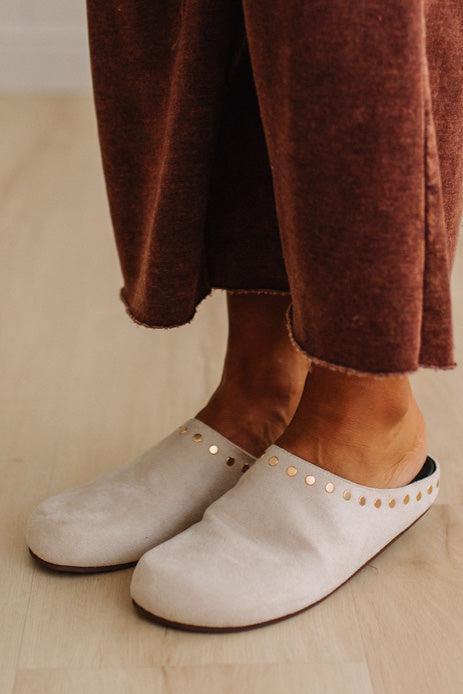 A close-up of a person wearing off-white slip-on clogs with gold-tone studs, paired with brown textured pants, standing on a light wood floor.