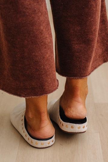 Close-up of a person wearing brown textured knit pants and off-white slide sandals with gold-tone circular studs on the sole, standing on a light wood floor.